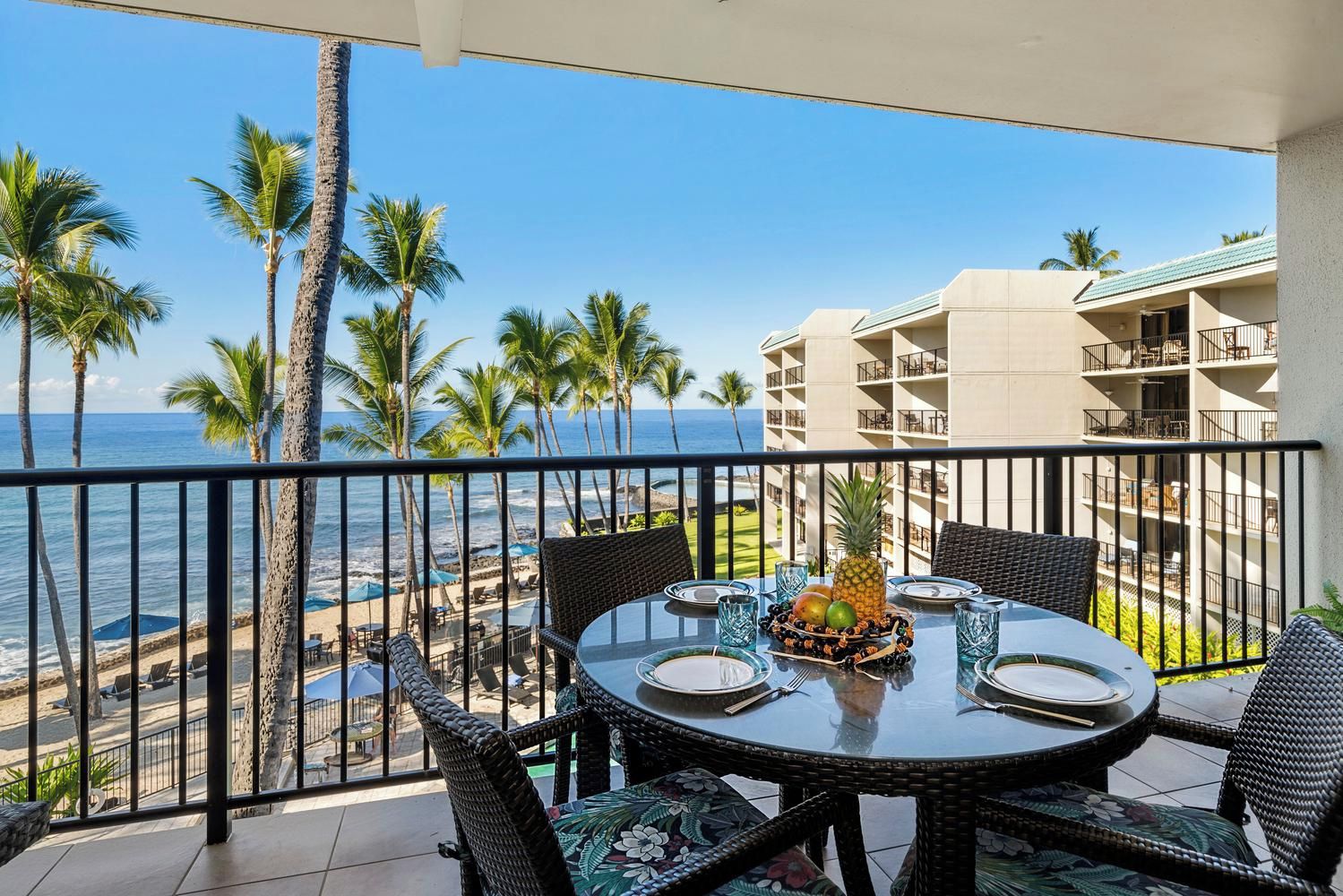 A balcony with a table set for dining, overlooking a beach with palm trees and a building in the background.