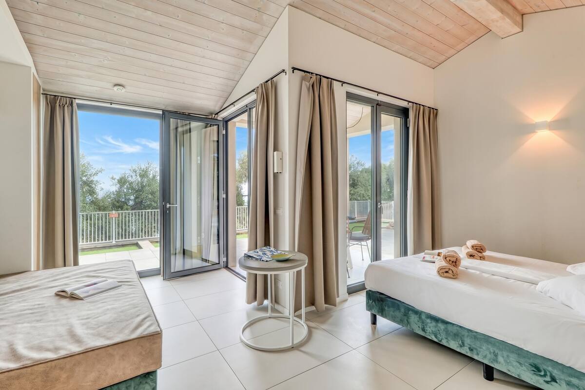 Une chambre d’hôtel avec un lit, une petite table, un balcon avec des rideaux et une vue sur la verdure.