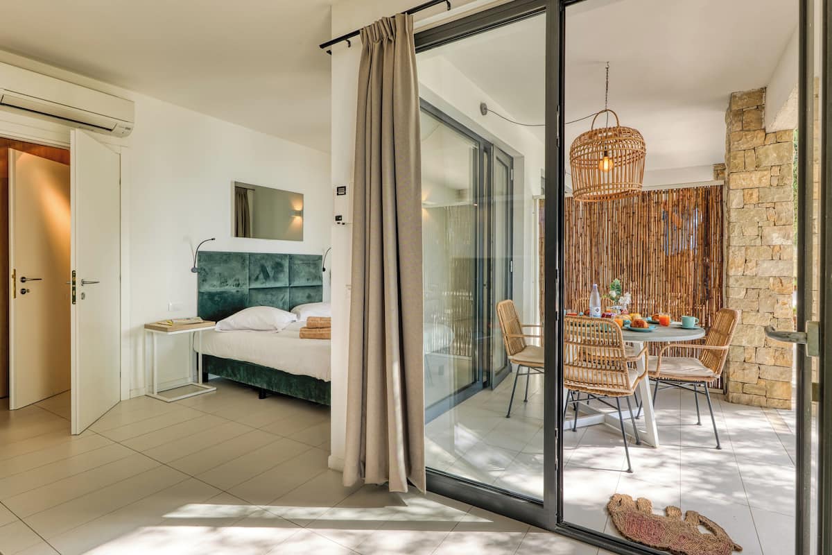 Une chambre moderne avec un lit, une table de chevet, une porte vitrée donnant sur un balcon aménagé avec des meubles en osier et un mur en pierre.
