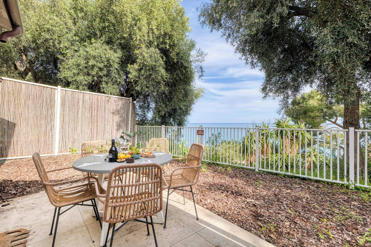 Une terrasse extérieure avec des chaises et une table en osier, donnant sur une étendue d’eau et entourée d’arbres.