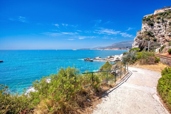Un sendero costero que lleva a un acantilado rocoso, con vistas despejadas al mar y una ciudad distante.