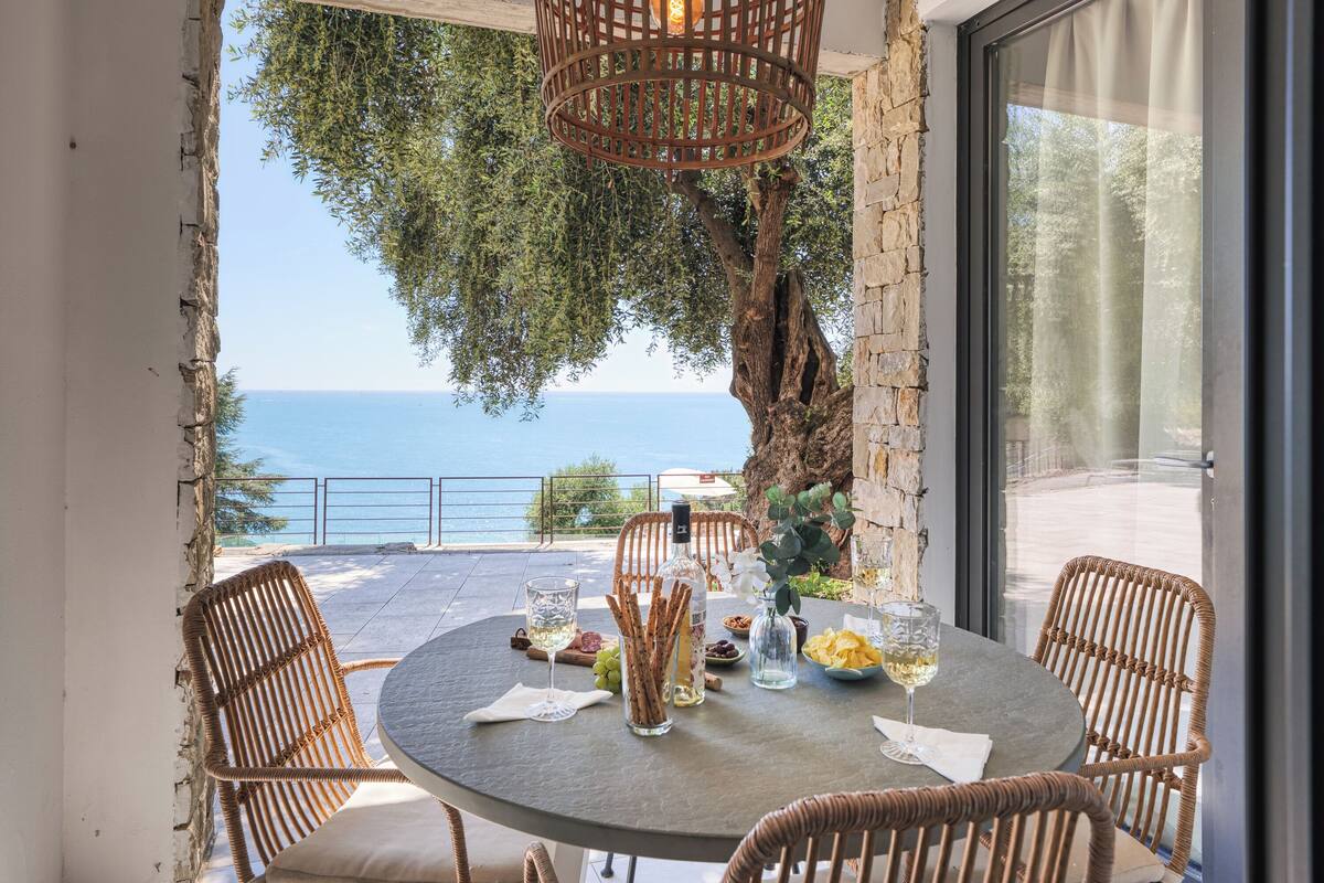 Une table ronde entourée de chaises en osier, une bouteille et des amuse-gueules, le tout sur un balcon donnant sur la mer.