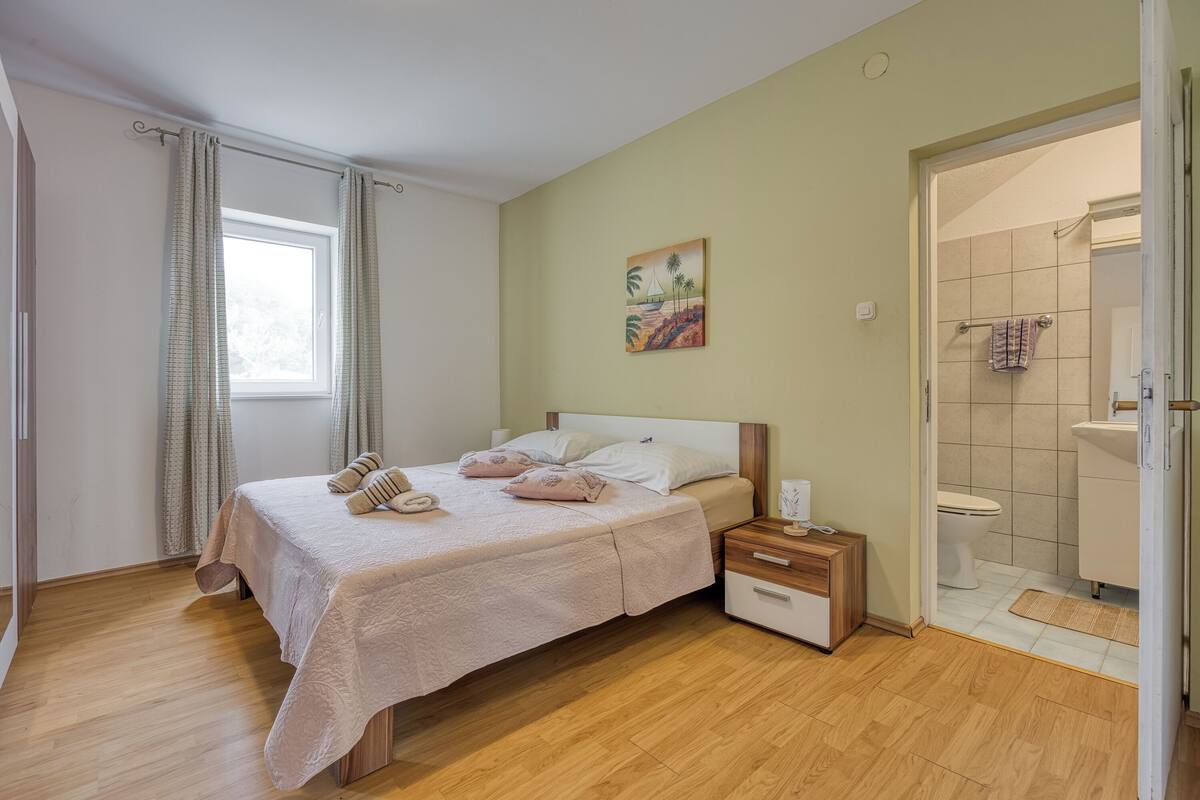 A bedroom with a bed, bedside tables, a window with curtains, a painting on the wall, and a bathroom with a toilet and sink.