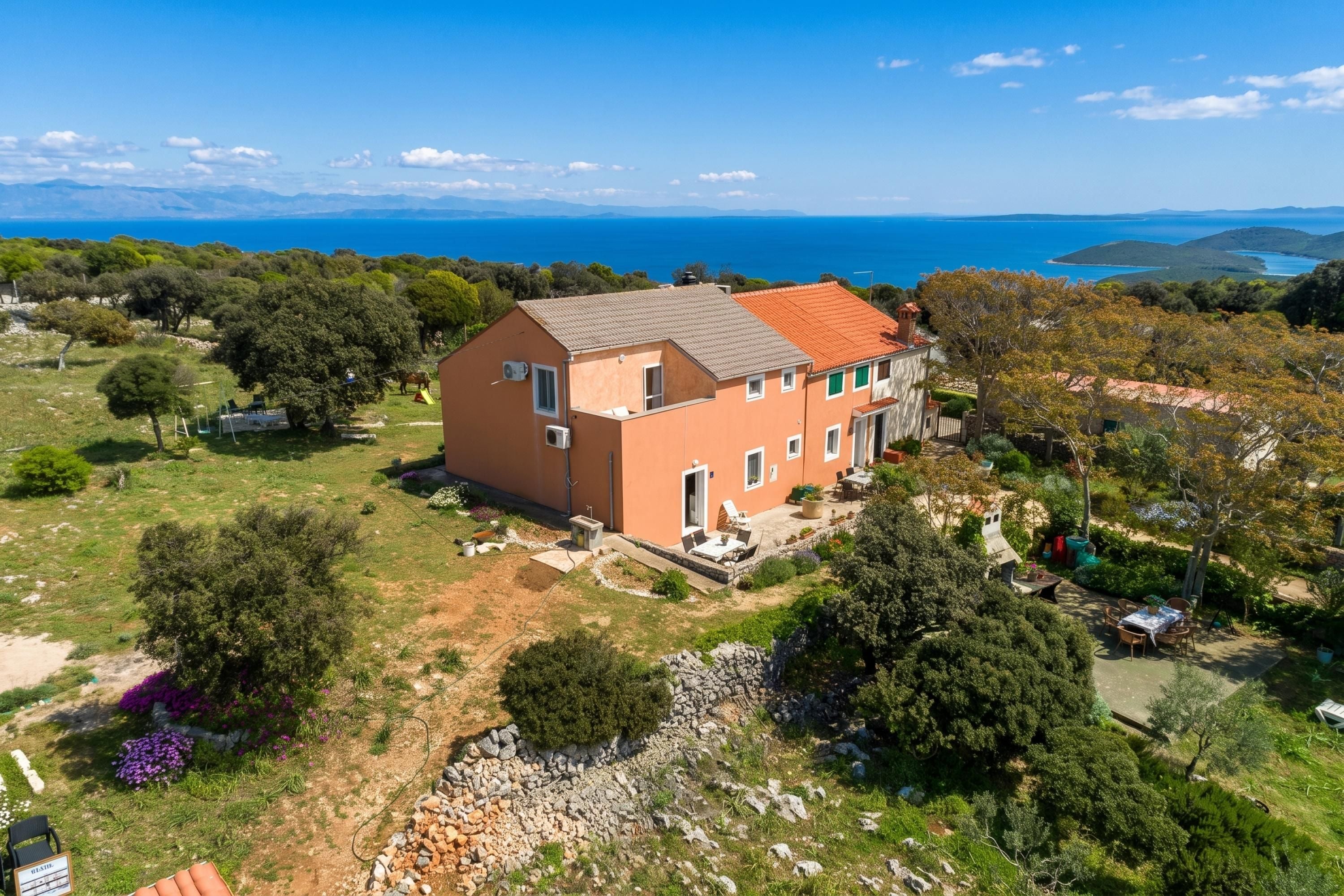 Uma casa de dois andares com telhado vermelho, rodeada por um jardim e vista para o mar.