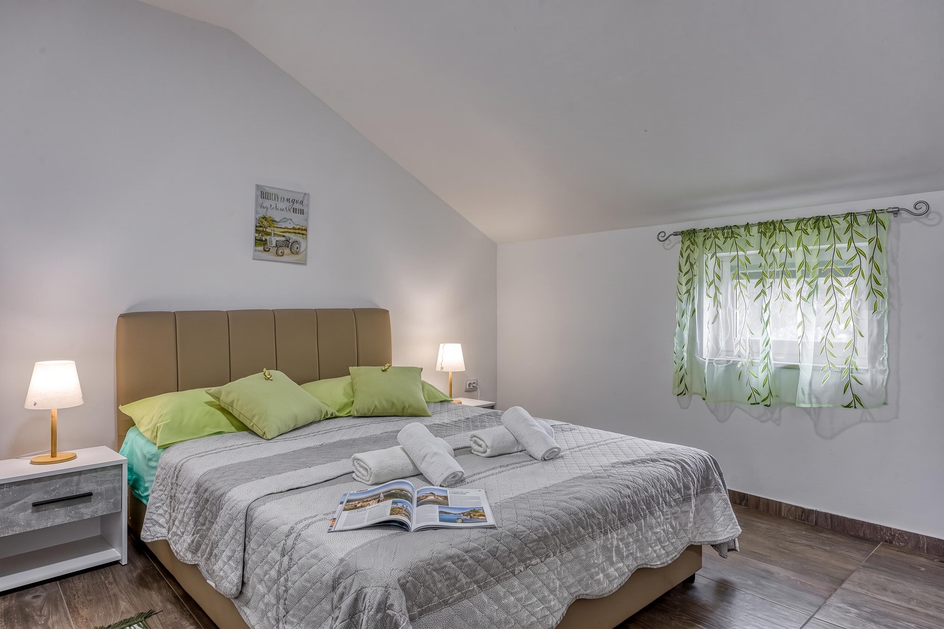 A bedroom with a bed, two bedside tables, a window with a curtain, and a wall-mounted picture.