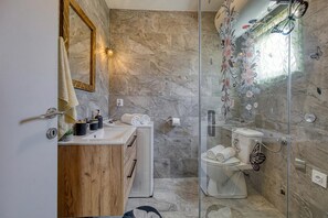 A bathroom with a marble wall, a wooden vanity, a glass shower enclosure, and a toilet with a butterfly decoration. - Island Top Sea View Estate (Mali Losinj)