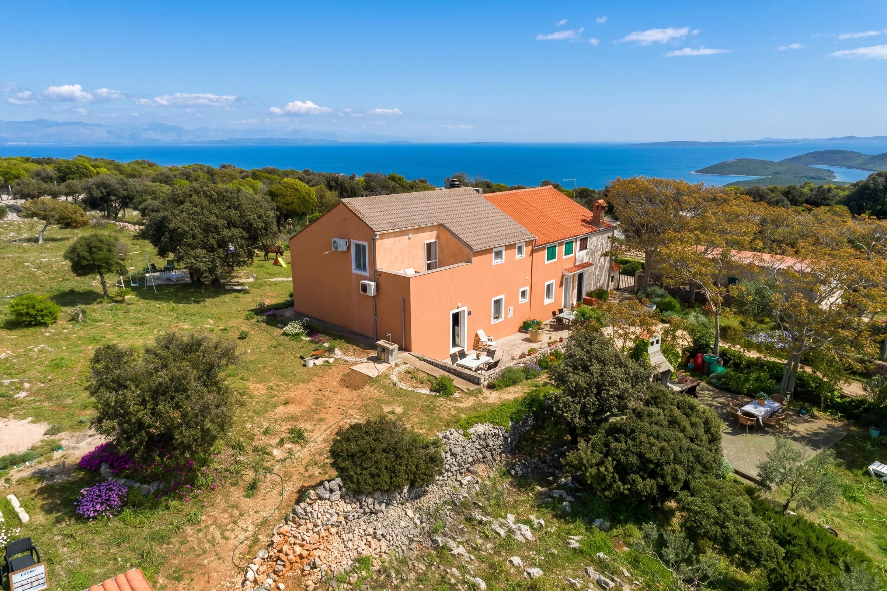 Una casa de dos pisos con tejado de tejas rojas, rodeada de un jardín y vista al mar.