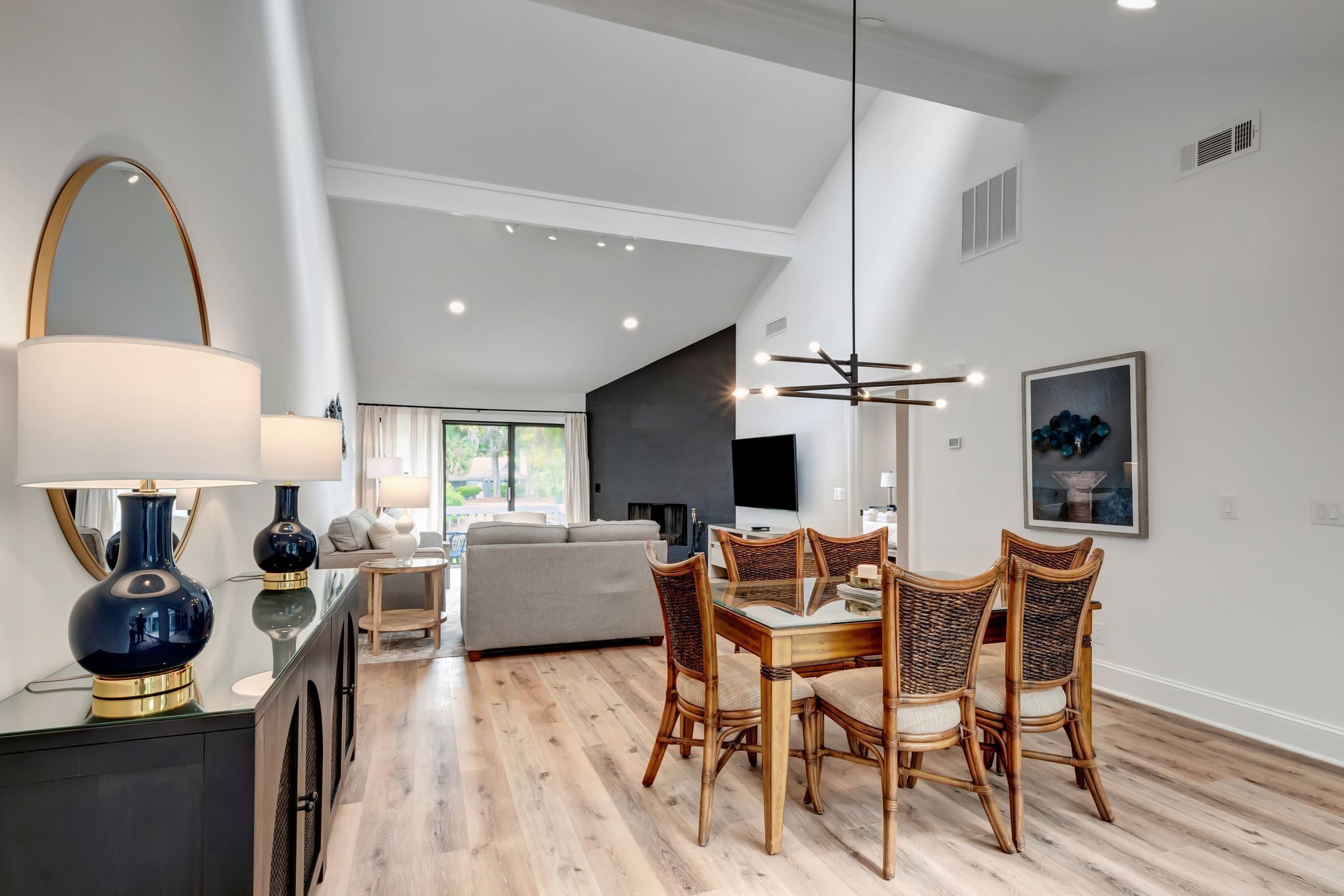 A modern dining area with a glass table and wicker chairs, a dark blue lamp, and a large mirror on the wall.