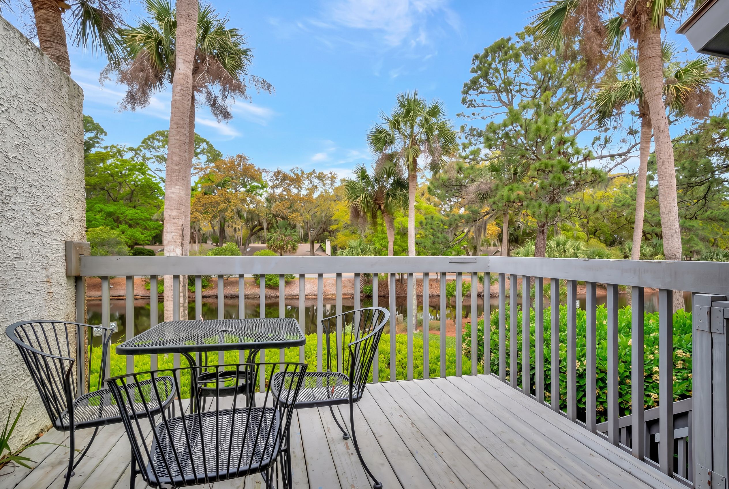 A wooden deck with a table and chairs, surrounded by a railing, overlooking a pond and various trees.