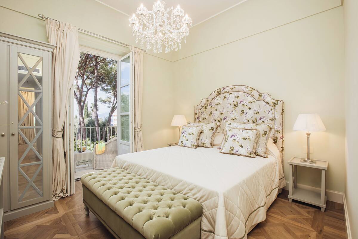 A bedroom with a large bed, a chandelier, a bench, and a view of the outdoors.