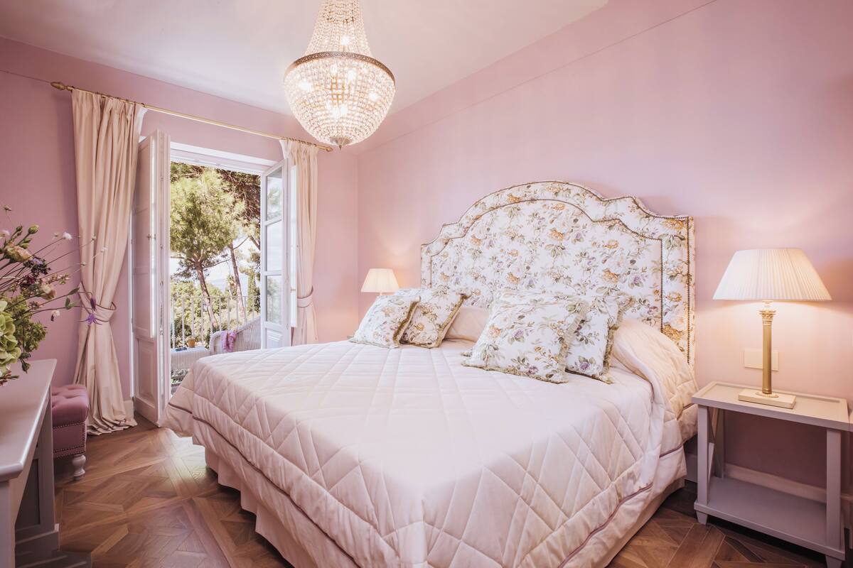 A bedroom with a large bed, two bedside lamps, a chandelier, and a view of the outdoors.