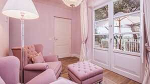 Una stanza in tonalità rosa con un lampadario, un divano, un poggiapiedi e una grande finestra che si affaccia su alberi.