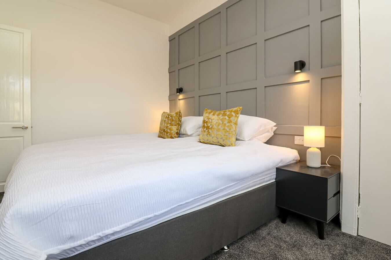 A neatly made bed with white bedding and two patterned pillows, a bedside table with a lamp, and a grey headboard.