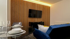 Una sala de estar moderna con una mesa de comedor de vidrio para dos personas, un sofá azul y un televisor de pantalla plana montado en una pared de madera.