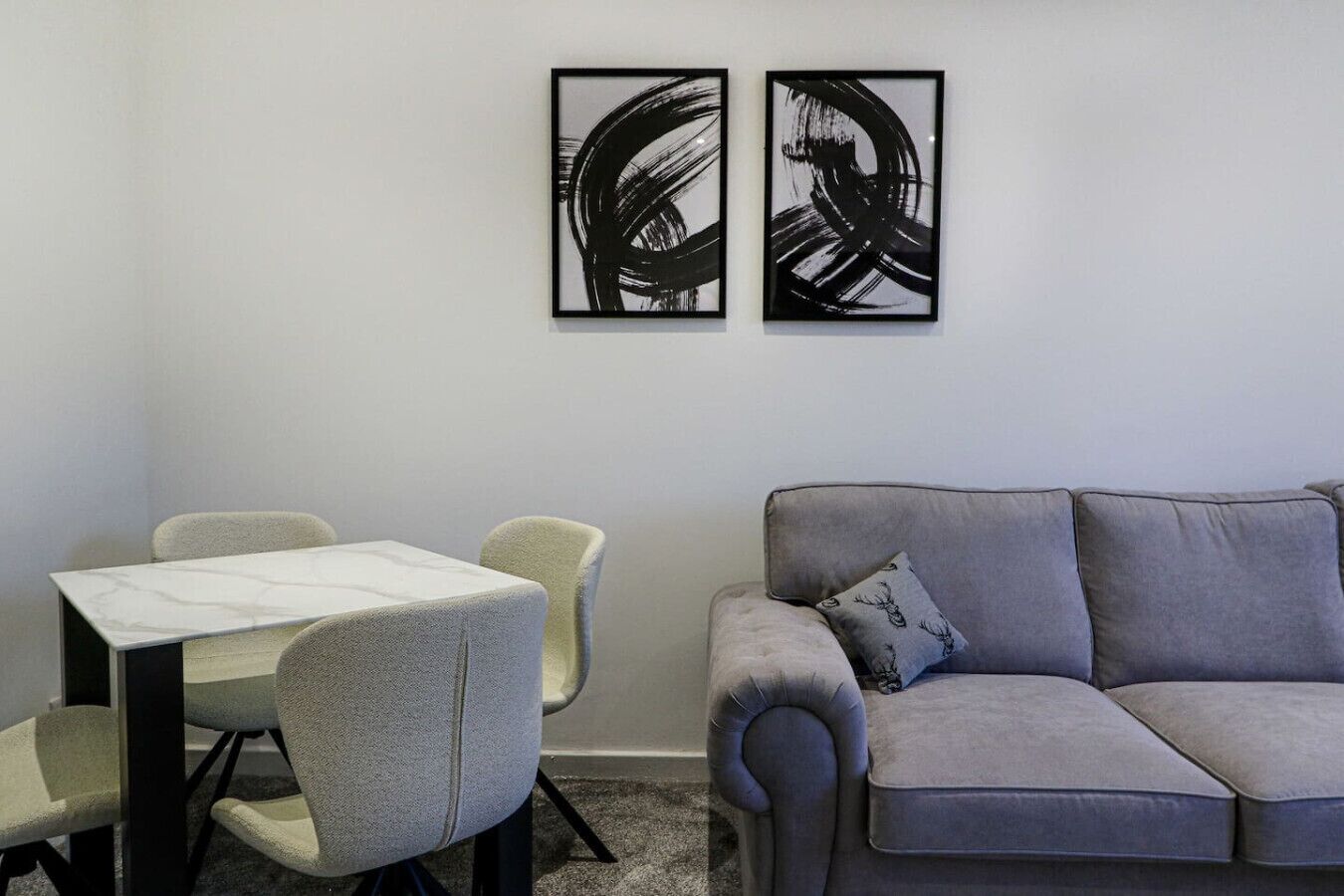 A modern living room with a grey sofa and a small table with two chairs. Two abstract paintings are hung on the wall above the sofa.