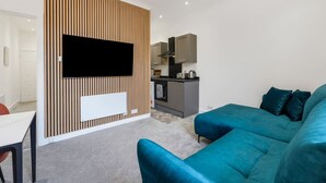 Una sala de estar moderna con un televisor de pantalla plana grande montado en una pared de paneles de madera, un sofá color aguamarina y una pequeña zona de cocina al fondo.