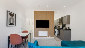Una habitación de hotel moderna con comedor, televisor montado en una pared de paneles de madera, kitchenette y cama con ropa de cama azul.