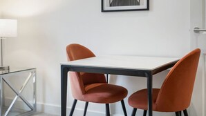 Un comedor moderno con una mesa blanca y sillas anaranjadas, una obra enmarcada en la pared y una división de vidrio a la izquierda.