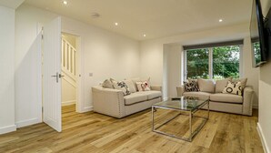 A spacious living room with a wooden floor, two beige sofas with patterned cushions, a glass coffee table, and a large window offering a view of greenery. - Beautiful 3 Bed Luxury House in Glasgow West End (Glasgow City)