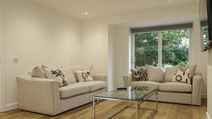 A modern living room with two beige sofas, a glass coffee table, wooden flooring, and a large window with a view of greenery. - Beautiful 3 Bed Luxury House in Glasgow West End (Glasgow City)