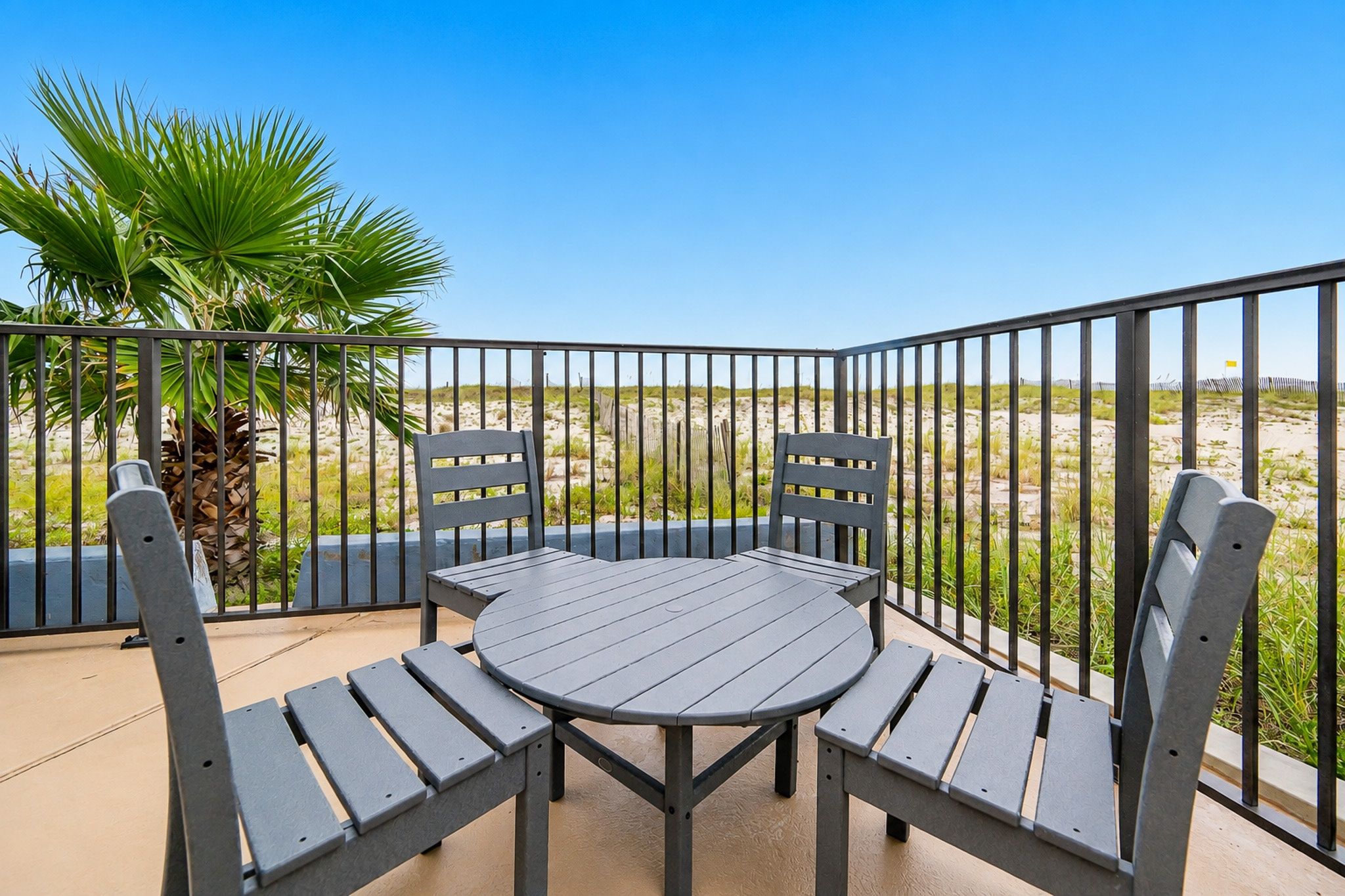 Ein Balkon mit rundem Tisch und vier Stühlen, umgeben von einem Metallgeländer, mit Blick auf einen Strand mit Dünen und spärlicher Vegetation.