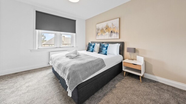 A bedroom with a bed, a nightstand, a lamp, a window with blinds, and a painting on the wall. - Cozy 2 bed apartment in quaint countryside (Lesmahagow)