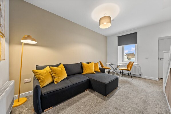 A modern living room with a sectional sofa, yellow cushions, a floor lamp, a dining table with chairs, and a view of the outside through a window.