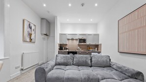 Una sala de estar moderna con un sofá seccional gris, una cocina con armarios blancos y un cuadro abstracto grande en la pared.