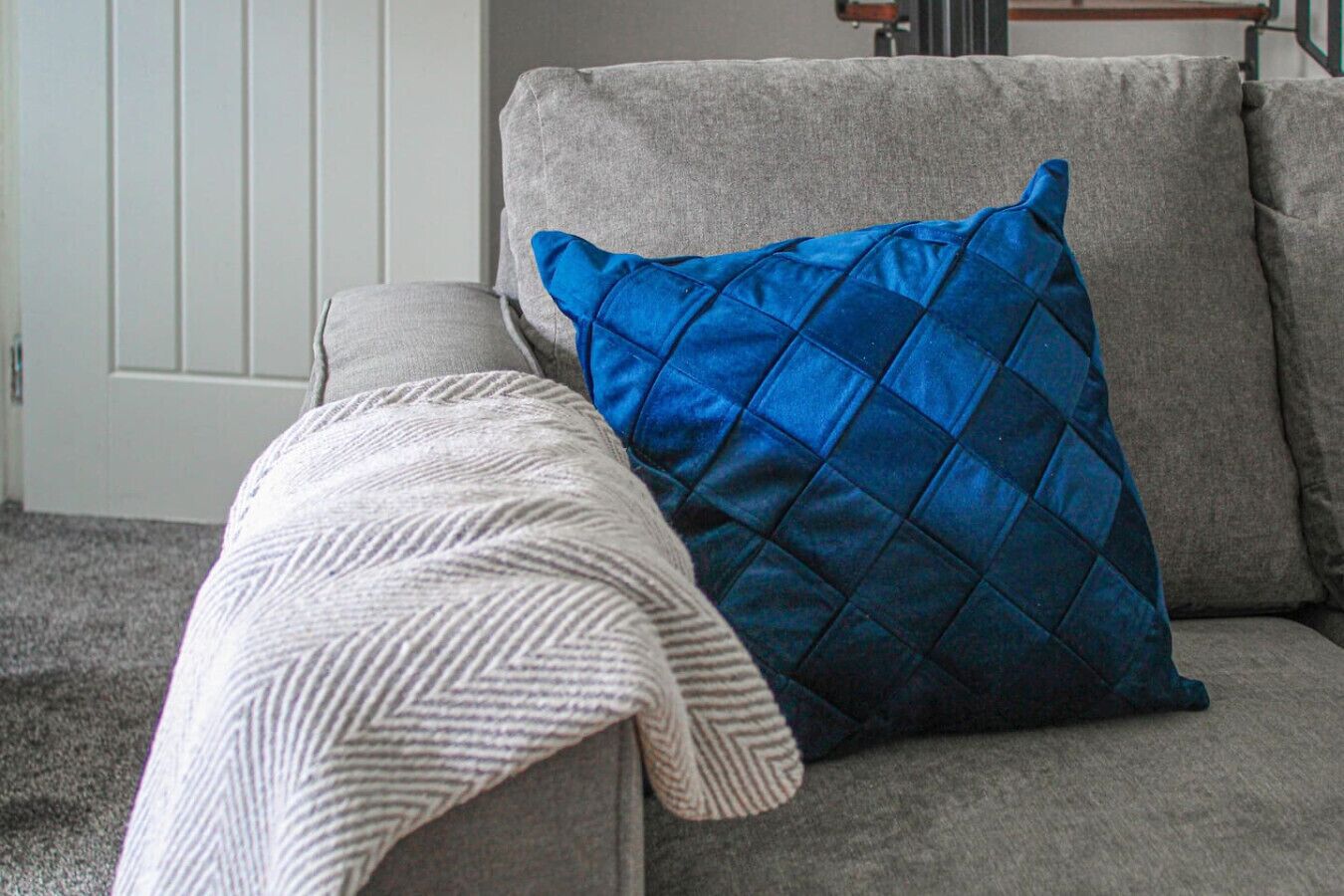A grey sofa with a blue quilted pillow and a white knitted throw.