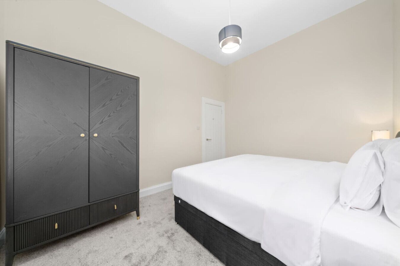 A bedroom with a large wardrobe, a bed with white bedding, a white door, and a ceiling light.
