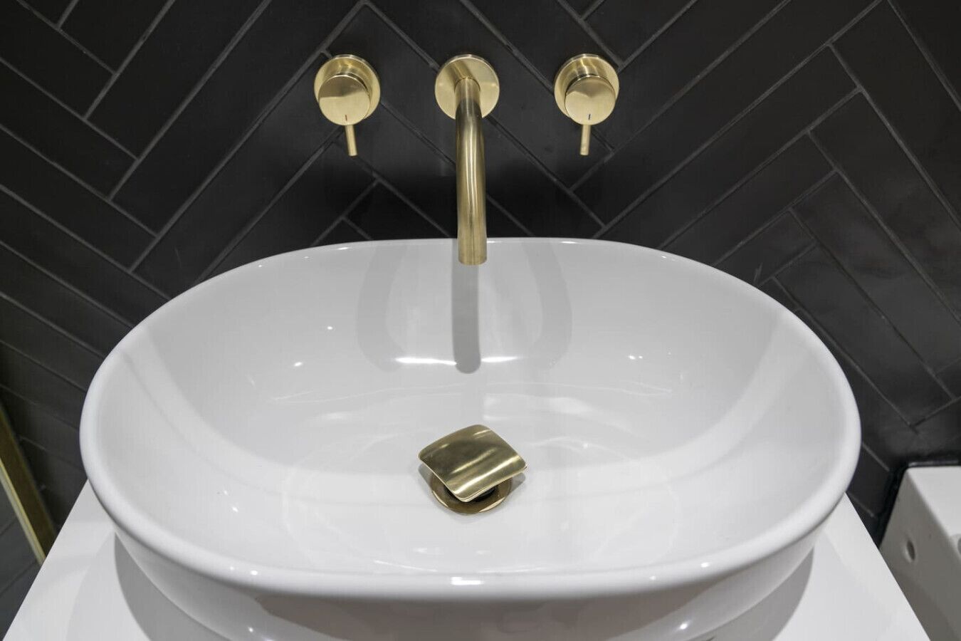 A white sink with a gold faucet and a gold soap dish in a bathroom with black tiled walls.