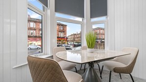 Un comedor moderno con una mesa de vidrio y cuatro sillas, ventanas grandes con vista a la calle y una planta en maceta sobre la mesa.