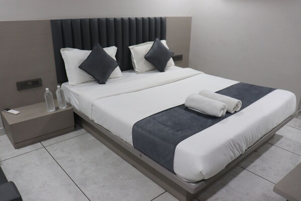 A modern hotel room with a large bed, a nightstand with a phone and water bottles, and a neatly made bed with towels. - Hotel R K villa (Ahmedabad)