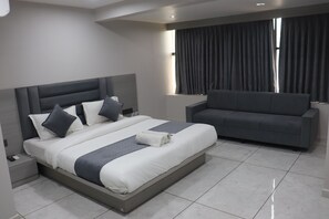 A modern bedroom with a large bed, a grey sofa, and a window with curtains. - Hotel R K villa (Ahmedabad)