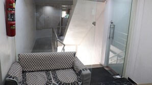 Eine Hotelhalle mit einer Couch, einem Feuerlöscher, einem gläsernen Aufzug und einer Glastür zu einem Duschbereich.