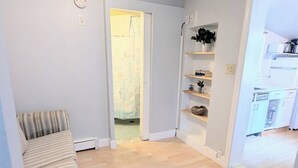 Habitació amb un sofà estampat, un terra de fusta, una porta que dona a un bany i una cuina visible a través d'una porta oberta.