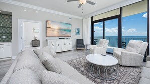 Ein geräumiges Wohnzimmer mit einer großen Couch, einem runden Couchtisch und zwei Sesseln. Ein Fernseher ist an der Wand montiert, und es gibt einen Blick auf den Ozean durch große Fenster.
