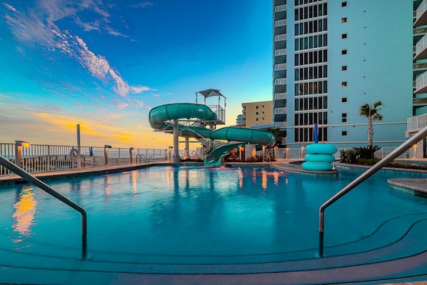 A swimming pool with a water slide, a tall building, and a sunset.