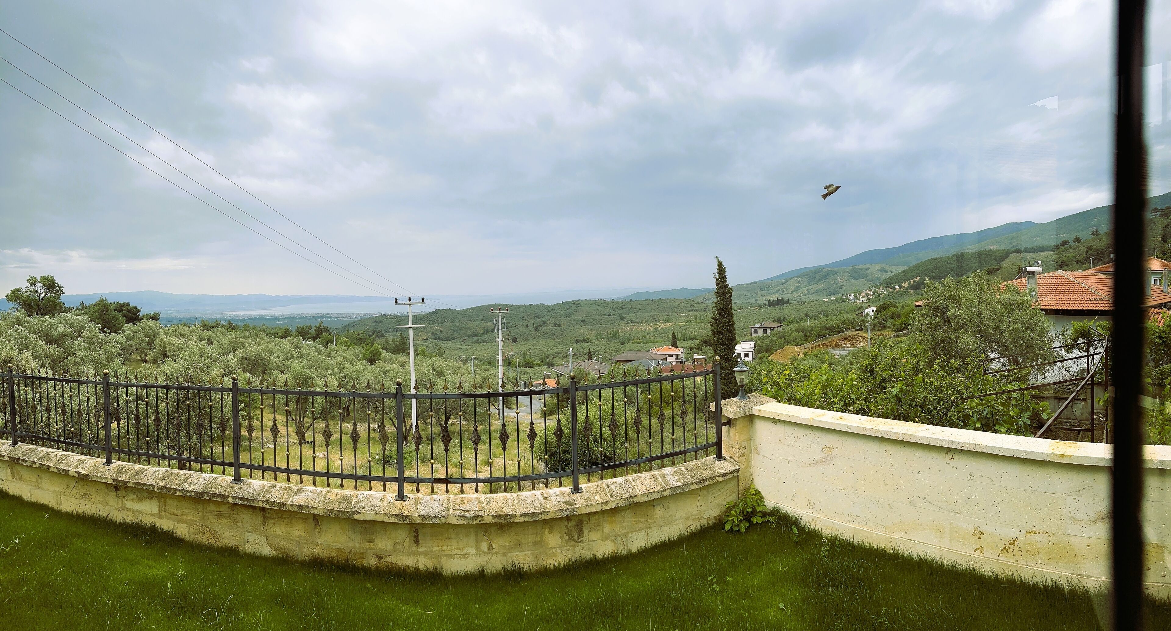 A view from a balcony overlooking a green landscape with a fence, a wall, and distant houses.
