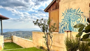 Uma área externa com gramado, uma cadeira de madeira, uma pequena mesa com um livro e um vaso de flores, e um edifício com um mural.