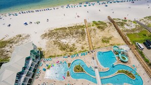An aerial view of a beach resort with a large swimming pool, beach umbrellas, and a wooden boardwalk.