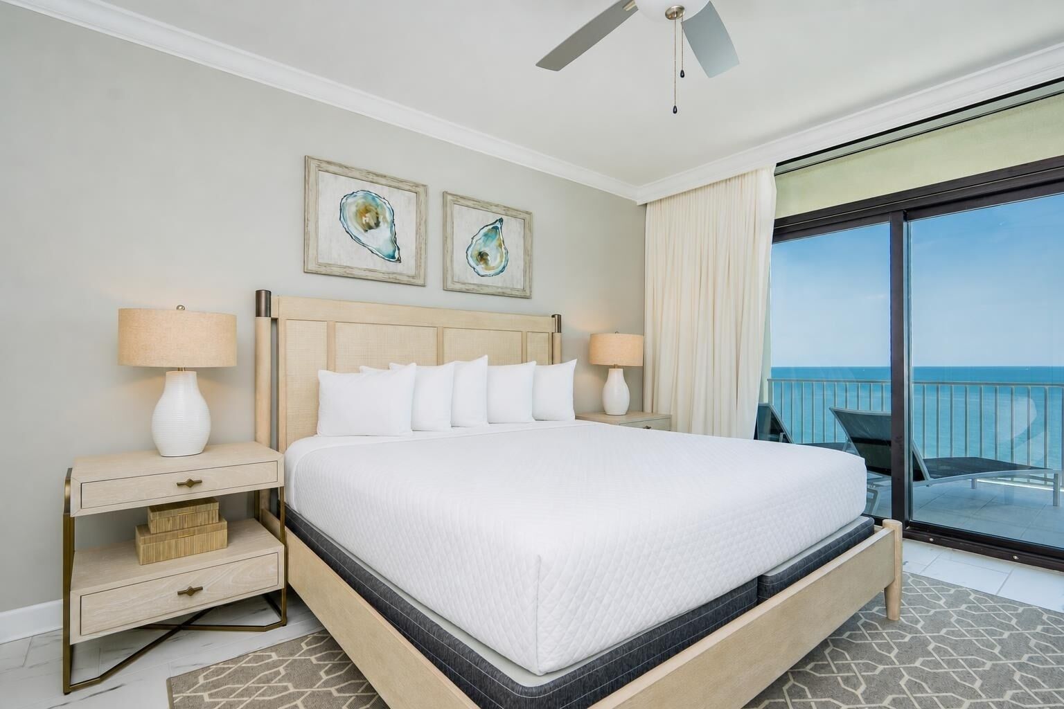 A hotel room with a large bed, two bedside tables with lamps, a ceiling fan, and a balcony with a view of the sea.