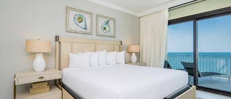 A hotel room with a large bed, two bedside tables with lamps, a ceiling fan, and a balcony with a view of the sea.