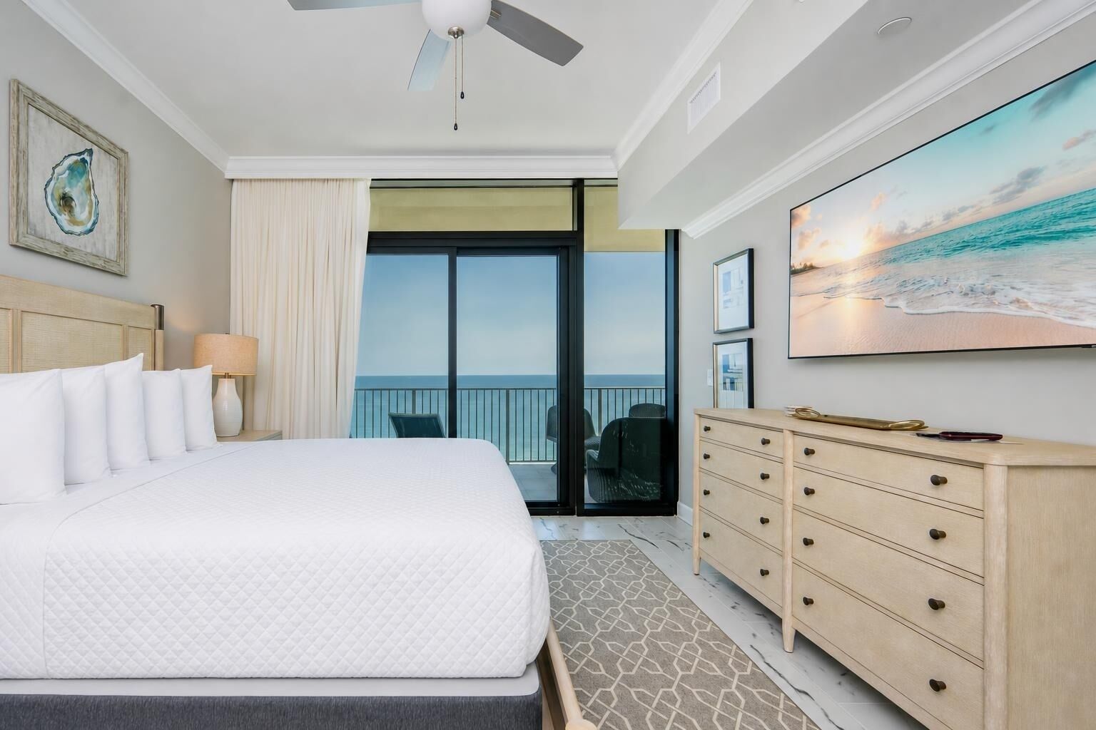 A hotel room with a bed, a dresser, a ceiling fan, a large window with a view of the ocean, and a painting on the wall.