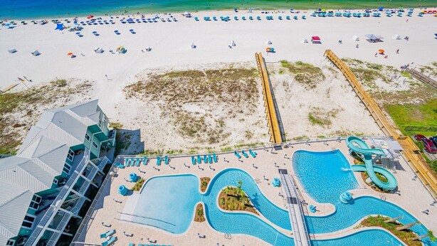 An aerial view of a beach resort with a large pool area, beach chairs, umbrellas, and a wooden boardwalk.