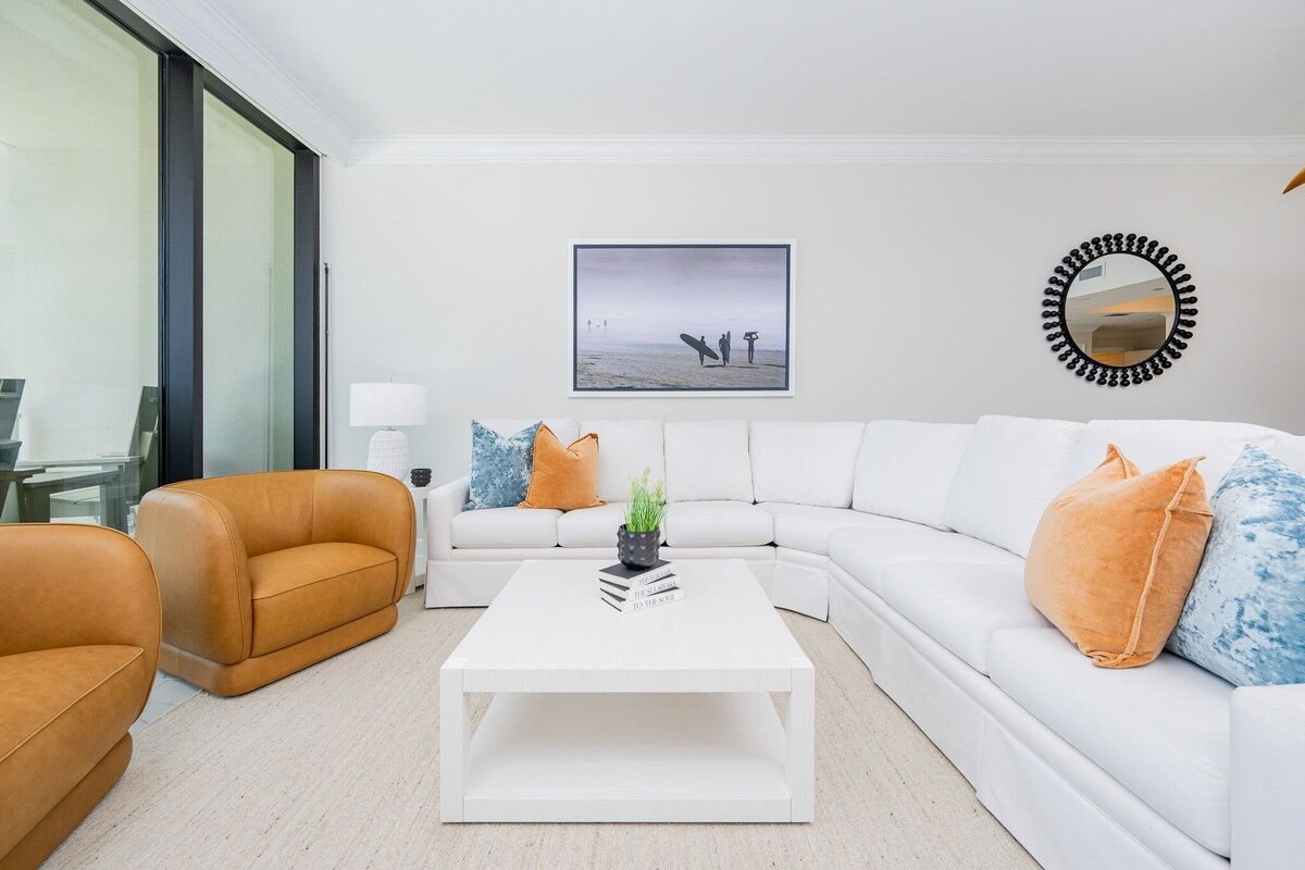 Un salon moderne avec un canapé blanc, un fauteuil en cuir marron et une table basse blanche. Un tableau encadré représentant une scène de plage est accroché au mur, et un miroir rond est placé au-dessus du canapé.