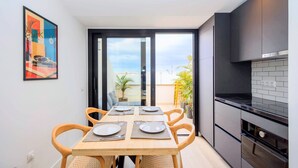 Una cocina moderna con armarios oscuros, una mesa de comedor preparada para cuatro personas y un amplio ventanal corredero que da a un balcón con vistas a la ciudad.