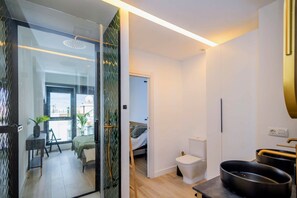 Een moderne slaapkamer met een glazen deur naar een balkon, een nachtkastje met een plant, een bed met hoofdeinde, en een badkamer met toilet en zwart wastafel.