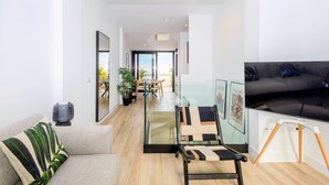 Una sala de estar moderna con un sofá, una mesa de centro de vidrio y un televisor. Al fondo, se observa un comedor con una mesa y sillas.