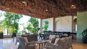 A spacious lounge area with wicker furniture, a thatched ceiling, and large windows offering views of greenery. - NEW LISTING** This premium two-bedroom condo offers the best of both worlds - set in one of Tulum’s most desirable areas, you’re just minutes from shops, restaurants, and the main road to the beach. (Tulum)